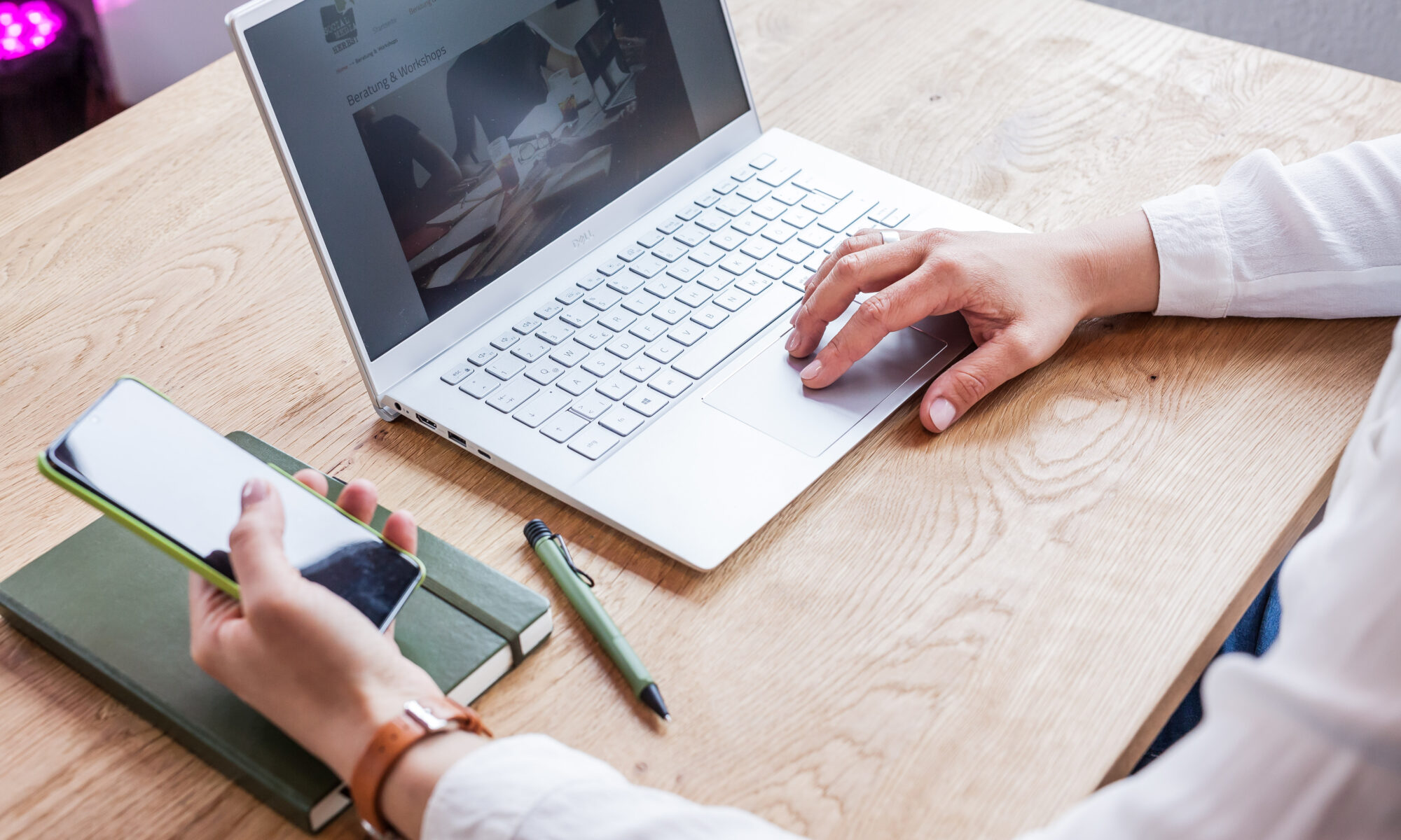 Auf einem Tisch steht ein Laptop, eine Person bedient ihn und hält in der anderen Hand ein Handy. Deneben liegt ein Notizbuch und ein Stift.